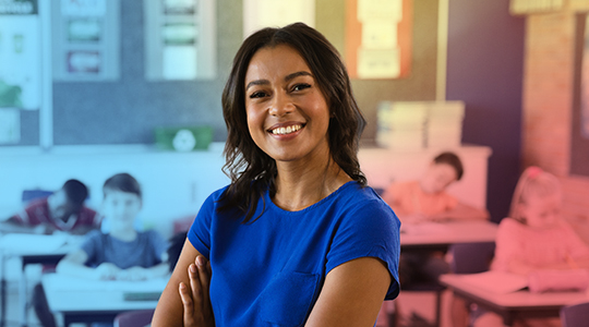 A smiling woman in a blue shirt stands with arms crossed in a classroom, with students working at desks in the background.