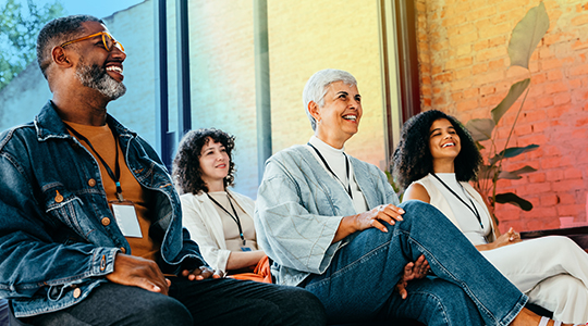 A diverse group of four adults wearing lanyards sit together, smiling and laughing in a warm, relaxed setting.