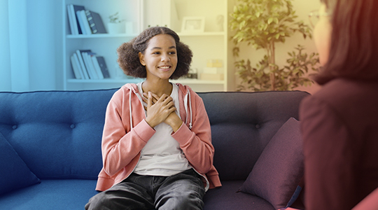 A teenage girl smiles and holds her hands to her chest while sitting on a couch, engaged in a positive conversation with an adult across from her