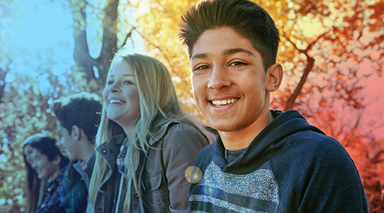 A smiling teenage boy stands outdoors with a group of peers laughing behind him among autumn trees