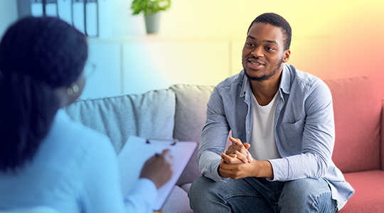 A young man sits on a couch and speaks openly with a professional who is taking notes, in what appears to be a counseling or therapy session