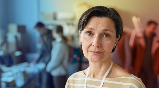 A woman wearing a lanyard looks calmly at the camera while volunteers work in the background, suggesting a disaster relief or community response setting
