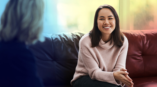A young woman smiles warmly while sitting on a couch across from another person, suggesting a supportive counseling or therapy session