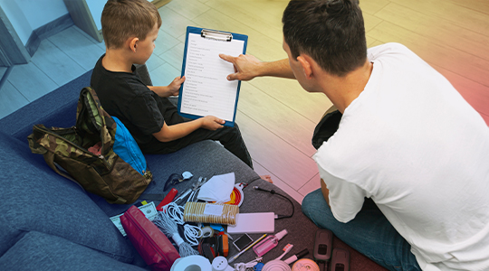 An adult and a child review a checklist on a clipboard together, with an emergency supply kit and backpack spread out in front of them