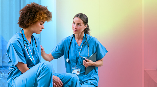 A healthcare worker places a supportive hand on the shoulder of a colleague in scrubs who appears distressed, while holding a clipboard