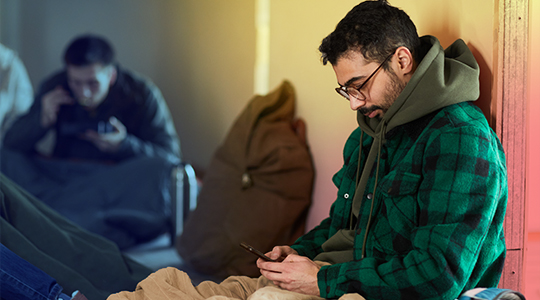 A young man wearing glasses sits alone against a wall, looking down at his phone in a dimly lit room, with another person visible in the background