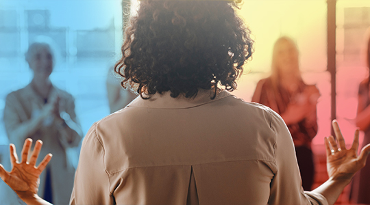 A woman with curly hair addresses a group of people, seen from behind with her hands raised, as the audience looks on attentively