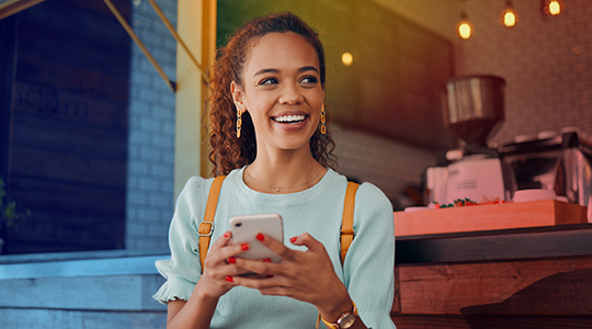 A young woman smiling and holding a smartphone outside a café, looking up with a confident and happy expression