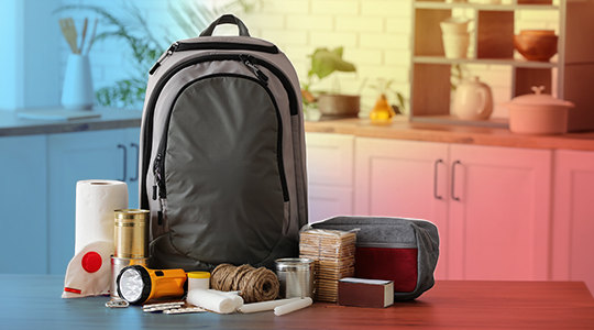 A disaster preparedness go-bag backpack on a kitchen table surrounded by emergency supplies including a flashlight, canned food, rope, bandages, matches, and paper towels