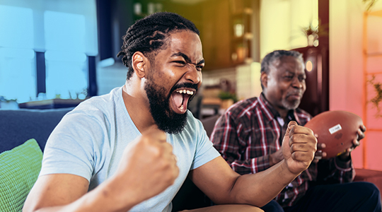 Senior father and adult son cheering together for an American football game at home